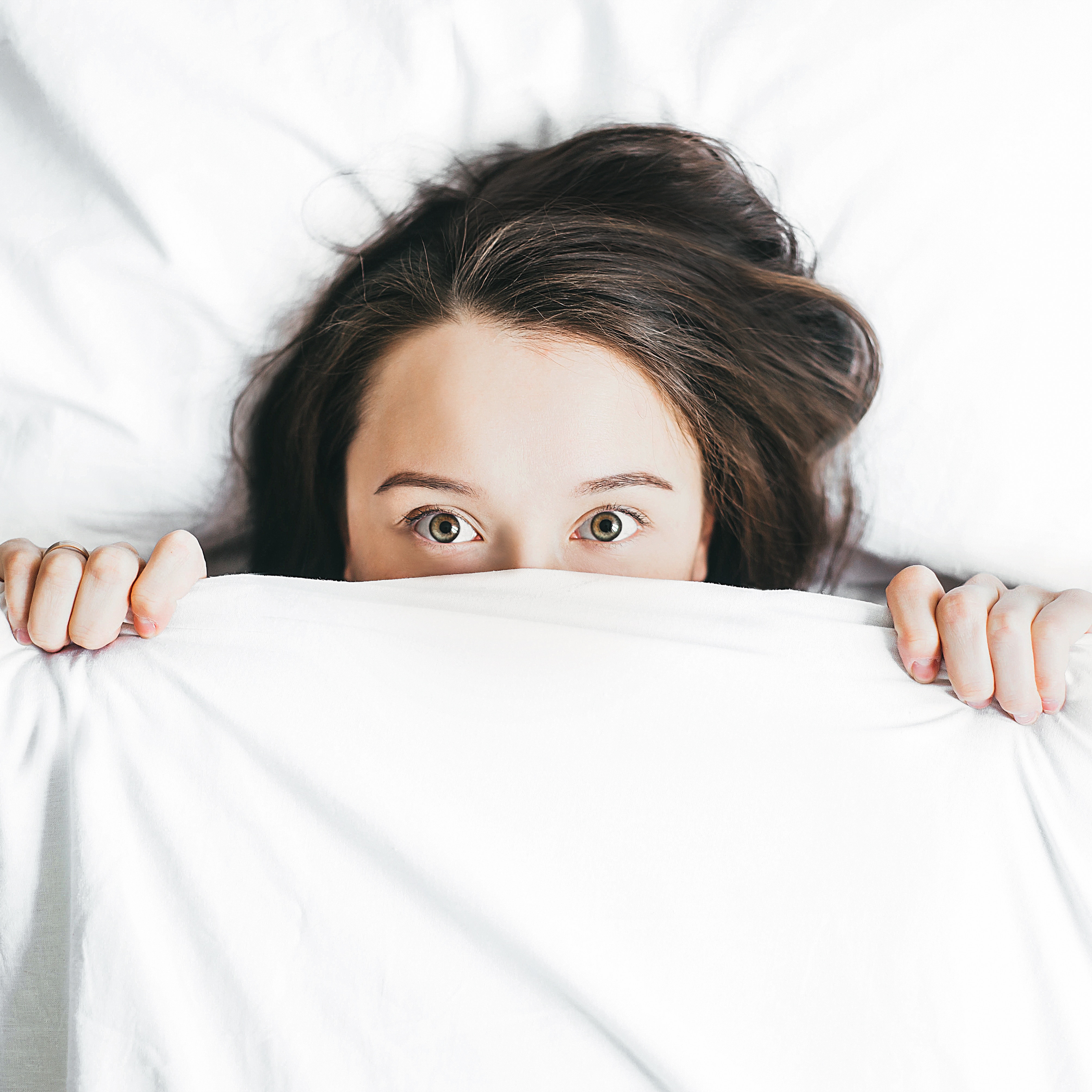 Young caucasian women with brown hair and hazel eyes lying in bed amongst white sheets hold the covers over her face until just under her eyes. She is wearing a 'I don't want to get out of bed' expression