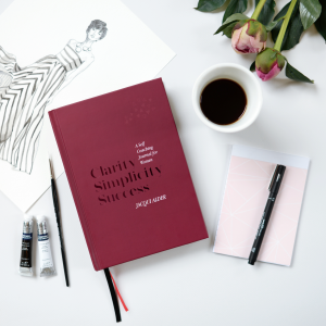 Burgundy coloured front cover of Clarity Simplicity Success A Self Coaching Journal for Women by Jacqui Alder on a desk with a beautiful fashion sketch and pink roses