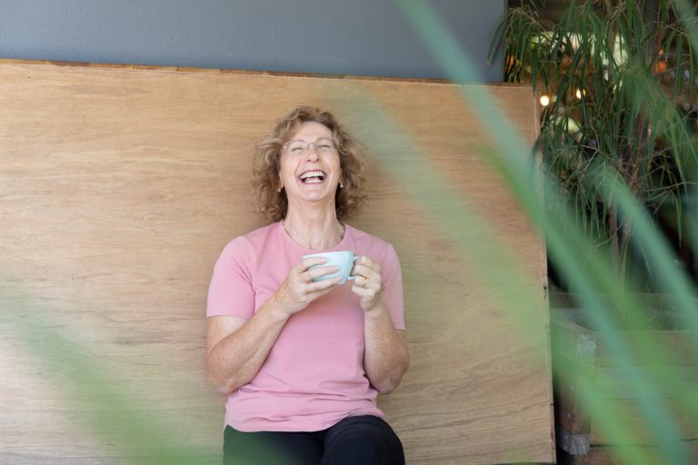 Woman (Jacqui Alder) laughing wearing pink top and holding a cup of coffee