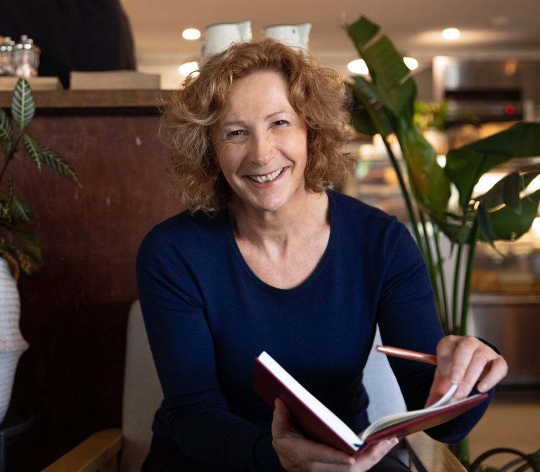 Smiling Jacqui Alder wearing blue top in a cafe with her book