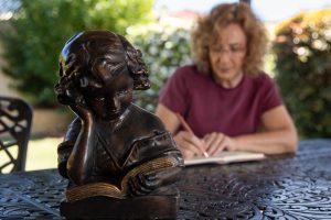 Jacqui Alder writing in a journal whilst seated at a table in her back garden. She us wearing a burgundy top. Figurine bust of girl reading in the left foreground.