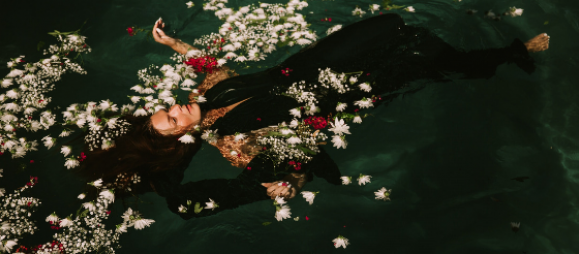 Dark green pool of water with woman wearing a black dress flaoting on on her back amongst floating white ad red flowers
